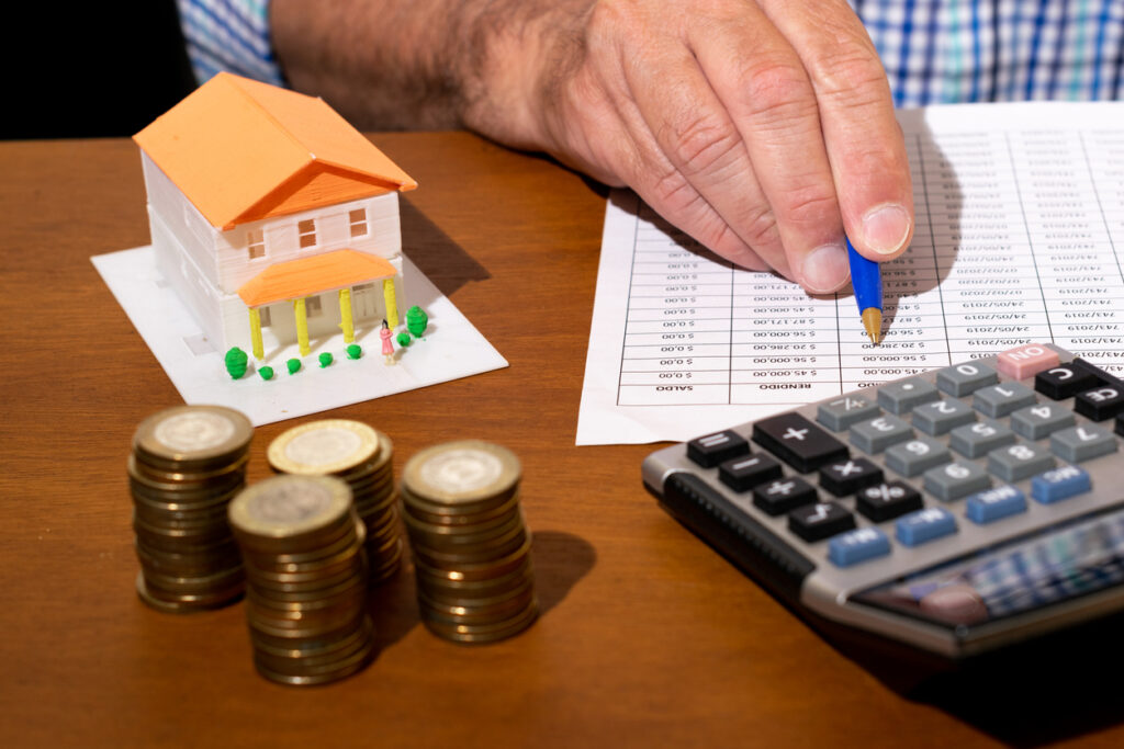 Partial view of person doing financial calculations. Coins in front, model of house and calculator